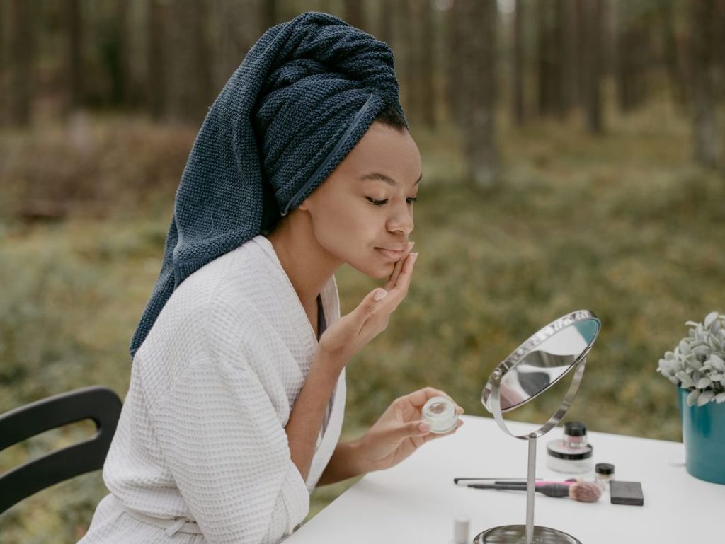 A woman in a bathrobe applies face cream in a serene outdoor environment.