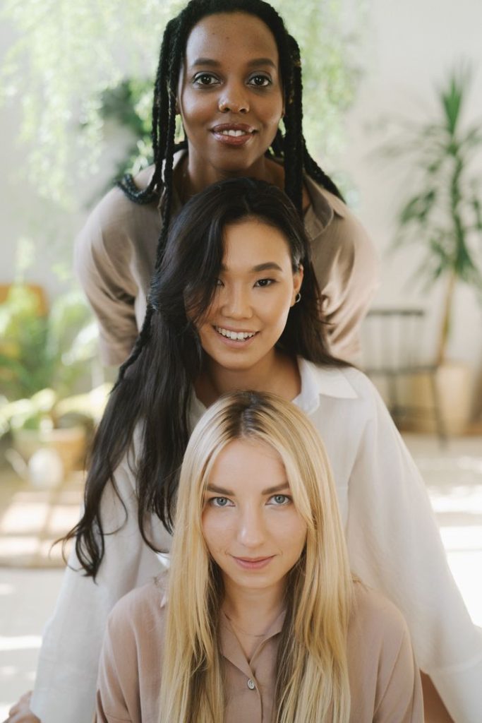 Portrait of three diverse women smiling indoors with natural lighting and greenery.