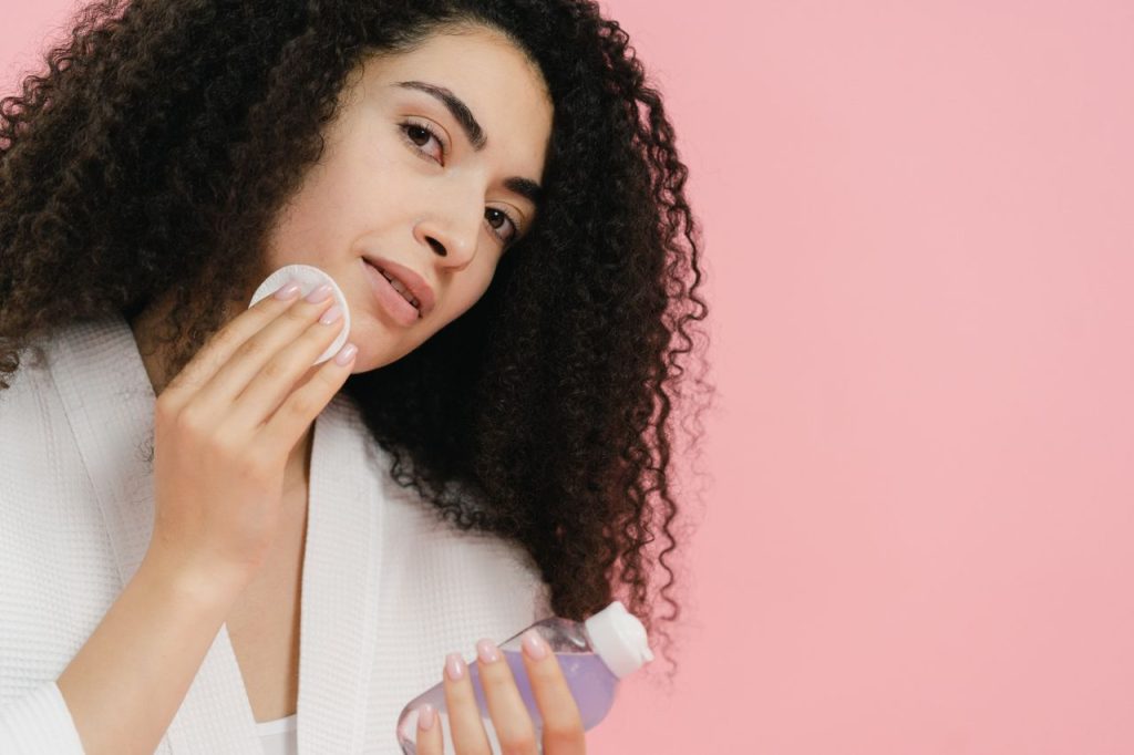 Young woman using a cosmetic pad and product for skincare against a pink backdrop.